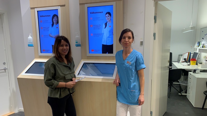Två mörkhåriga kvinnor, kvinnan till höger vårdpersonalklädd i blå blus och vita byxor,  står framför två digitala stationer med stora upplysta skärmar med texten Välkommen, som hänger på väggen över skrivdisplayer i midjehöjd.
