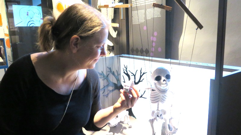 Blond kvinna i svart tröja framför liten marionettscen med ett skelett.