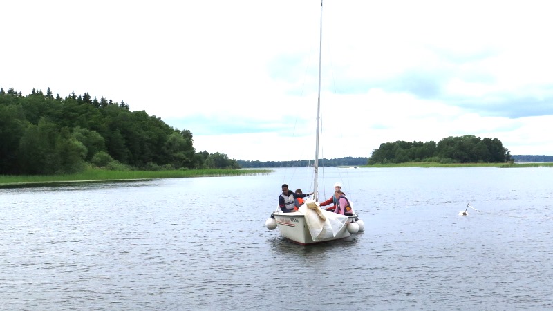 Fyra personer i flytväst i en mindre segelbåt på avstånd, omgivna av vatten, molnig himmel och grönskande öar.