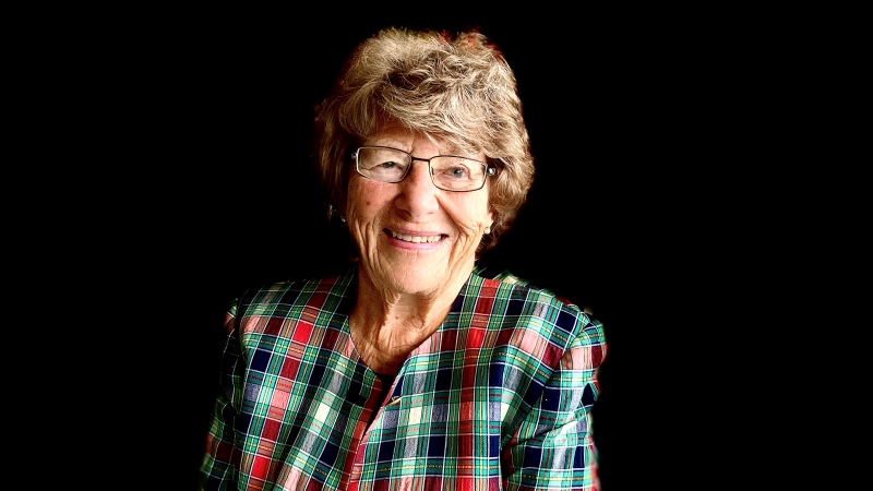 Ann-Mari Friman Persson, en äldre vit kvinna med kort lockigt grått hår, i grönrödrutig kavaj. Svart bakgrund.