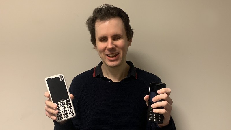  En vit man med halvlångt vågigt hår i mörkblå tröja ler och håller en mobiltelefon i vardera handen, en är vit och en svart.