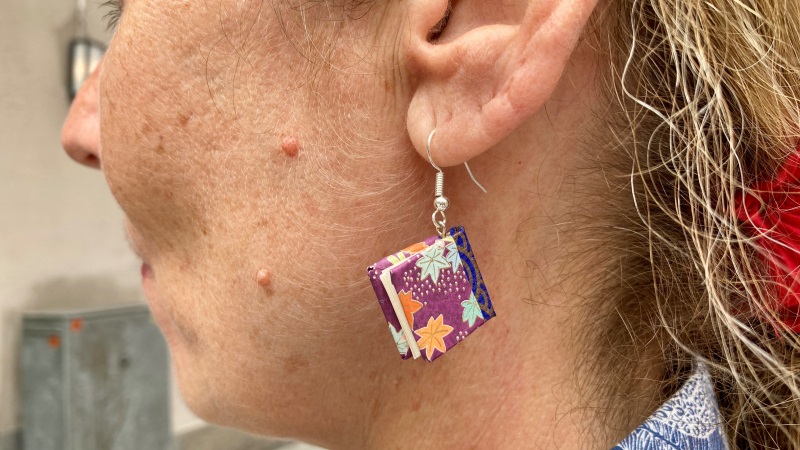 Närbild på ett örhänge som hänger i en kvinnas öra. Hänget har formen av en inbunden bok i miniatyr, med färgglatt omslag med mönster av sjöstjärnor.