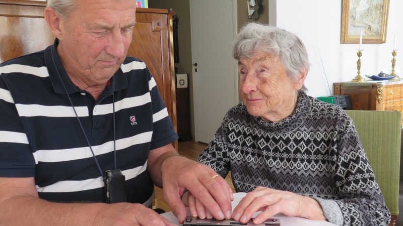 En gammal vit kvinna med grått hår, sitter vid köksbord med äldre vit man. Båda håller händer på en punktdislplay