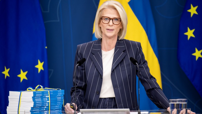 Finansministern, en blond kvinna med rakt hår och glasögon med sköldpaddsmönstrade bågar. Här i kritstrecksrandig kostym, enkel vit blus och med budgetluntan inbunden i blågult, framför svenska flaggor och EU-flaggor.