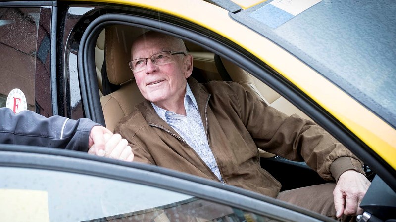 En ljus äldre man, sitter i en taxibil med dörren öppen. Det är färdtjänst.