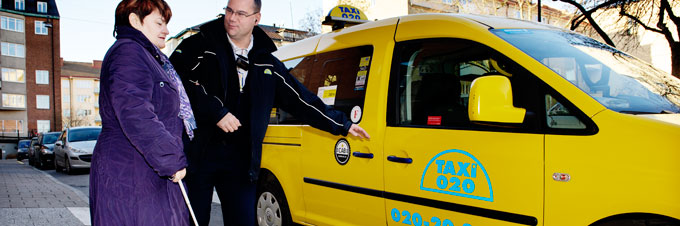 En kvinna med vit käpp är på väg till en gul taxibil, chauffören intill henne sträcker sig för att öppna dörren.