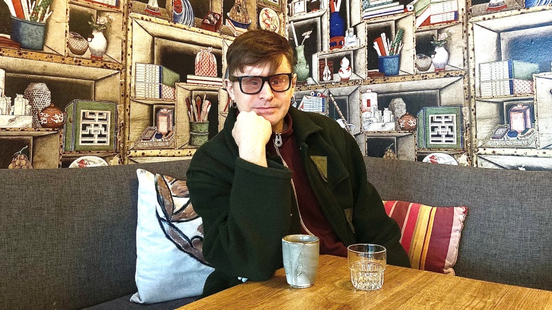 En medelålders vit man, kort brunt hår, glasögon, brun jacka, sitter vid träbord på café. En tapet med tavelramar i bakgrund