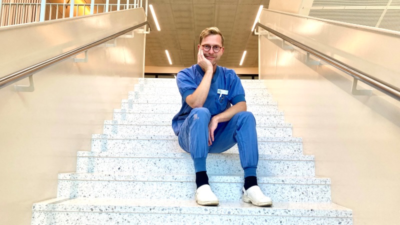 Blond, vit 40-årig man, blåa sjukhuskläder, sitter i vitt trapphus.