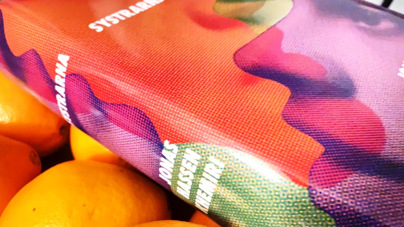 En tjock bok ligger på ett par apelsiner. Omslaget är ansikten i orange, lila och grönt. Systrarna - Jonas Hassen Khemiri.