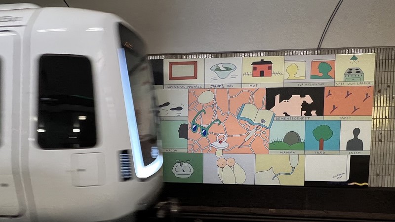 Ett suddigt tunnelbanetåg i rörelse till vänster. På väggen bakom syns konsten, bildrutor med en illustrerad tanke i varje, och ord. Till exempel: Hus, spis och lampa, träd, mamma (visar en gravsten).