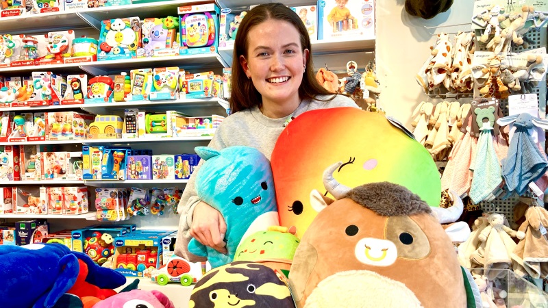 Rebecca Sundin, butikschef World of Toys är ung med ljust hy, långt brunt hår. Hon håller färgglada runda sagofigurkuddar.