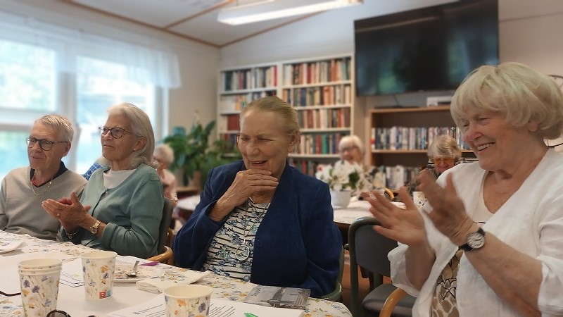  Ett gäng SRF-medlemmar är samlade runt ett blomsterprytt bord, och skrattar och applåderar. Längst till höger Maritta Söderholm, trivselansvarig i SRF Norrort.
