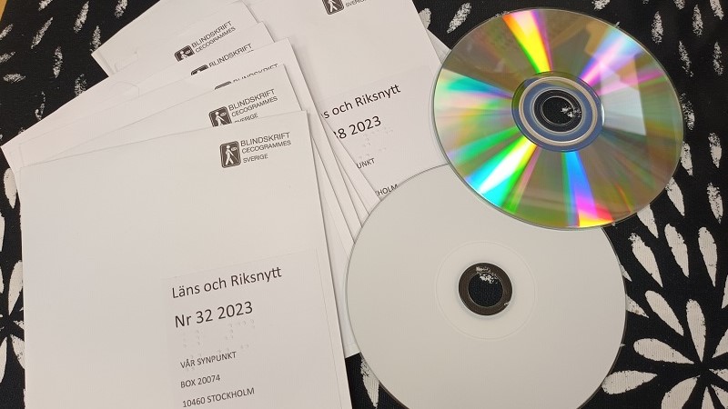 En hög med cd-skivor i kuvert på ett svartvitt tyg. Två skivor är utan kuvert, den ena vit och den andra regnbågsblank, med undersidan upp.