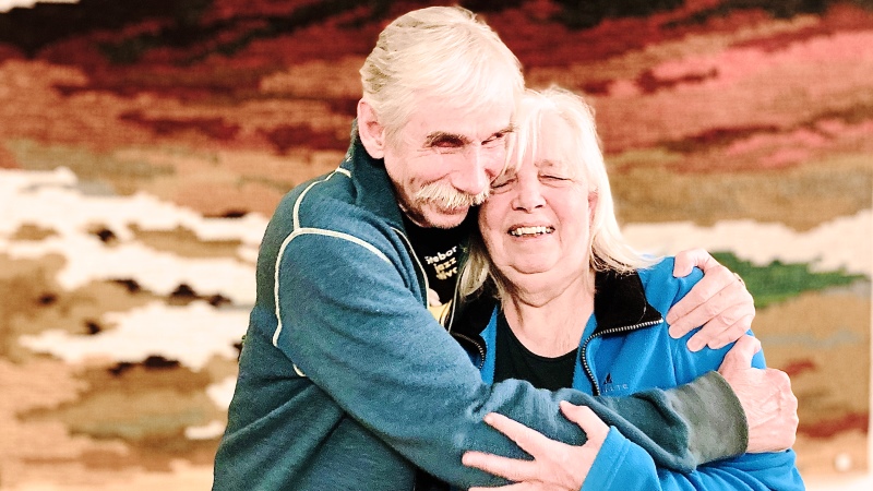 En man och en kvinna omfamnar varandra och ler. Båda är vita och har vitt hår, och är klädda i blått. I bakgrunden en väggtextil i jordfärger.