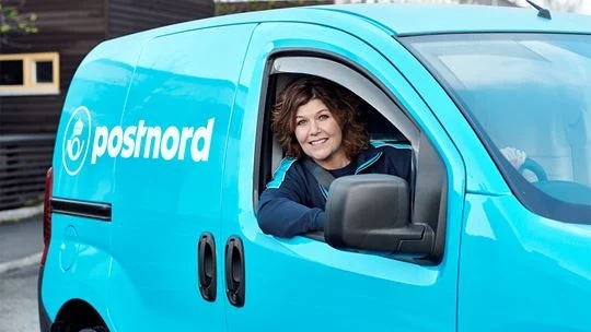 En leende kvinna tittar ut från förarsätet av en bil märkt med Postnord.