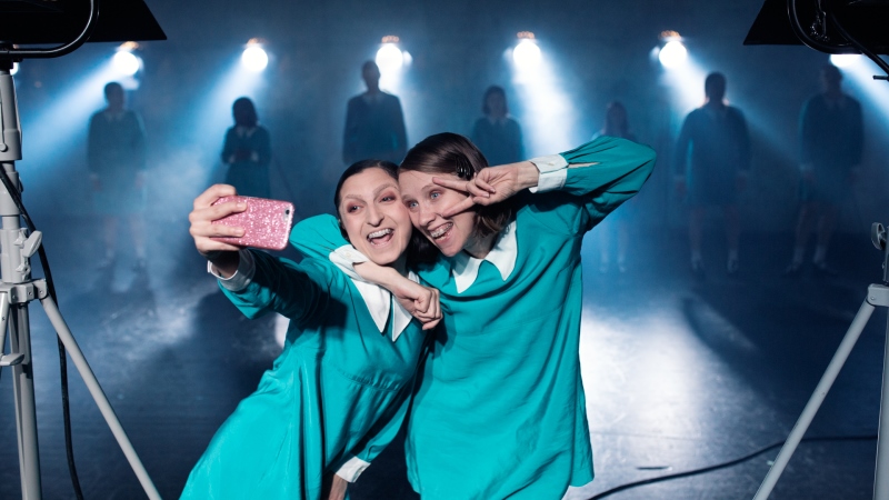: Två kvinnor i likadana turkosa skoluniformsklänningar tar en selfie med mobilen. Bakgrund ett mörkt teaterscenrum.