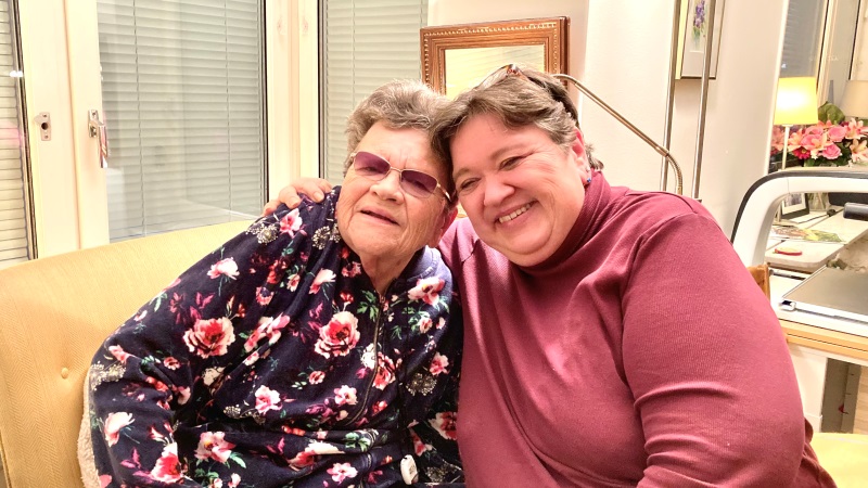 Två vita kvinnor sitter bredvid varandra i ett vardagsrum. Den ena är 80 år, den andra medelålders. De håller om varandra.