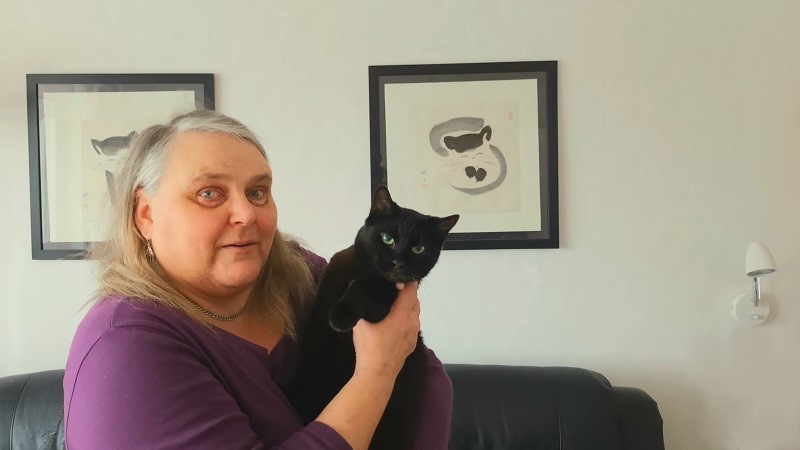 En kvinna med långt blont hår med gråa stråk håller en svart katt i famnen. Hon står i vardagsrummet vid en svart skinnsoffa och två målningar med kattmotiv.