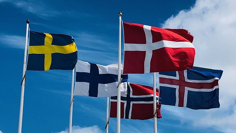  De fem nordiska ländernas flaggor vajar mot en blå himmel. Sverige, Danmark högst i bild närmast kameran. Därefter Finland och Island under och bakom. Längst bort i mitten är Norges flagga.