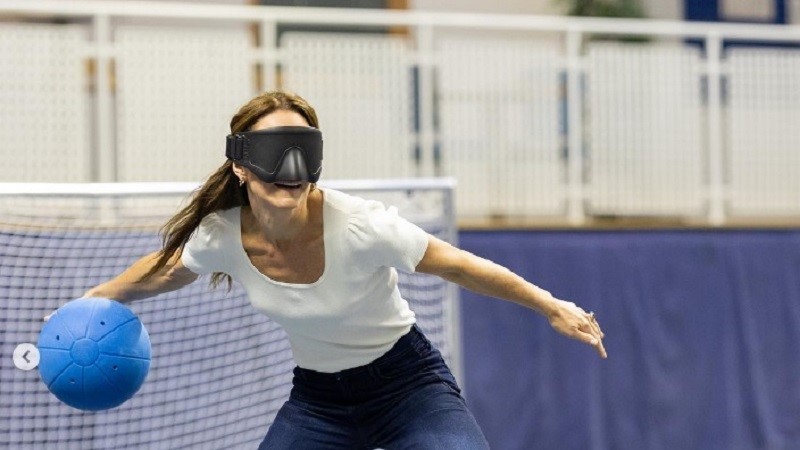 Kate Middleton med en blå goalball-boll i handen och armarna utsträckta åt varsitt håll på väg att göra ett kast. Hon har svart ögonbindel, hästsvans, blå stretchjeans och en vit puffärmad top.