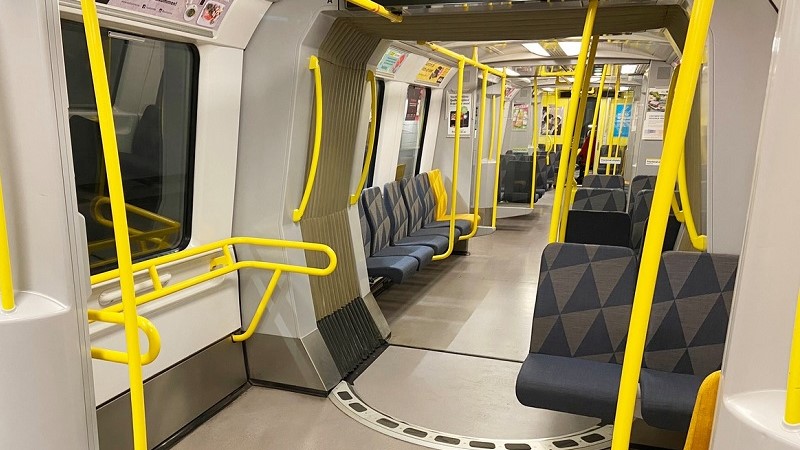 Insidan av en nyrenoverad tunnelbanevagn. Gula ledstänger kantar bilden och ett säte för två personer i grått tyg syns till höger.