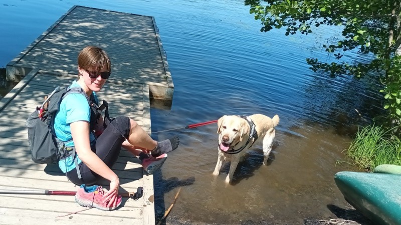 En ung kvinna i löpkläder och lätt ryggsäck sitter på en brygga. I vattnet står hennes ledarhund och flämtar.