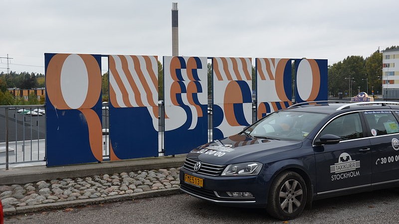 En taxibil står till höger i bild framför konstverket "Hej patient" vid entrén till Huddige sjukhus. Verket består av paneler med blått, vitt och aprikosfärgade former på.