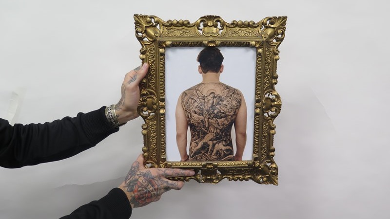 Två tatuerade händer håller mot vit bakgrund fram en guldram med foto av en heltatuerad rygg.