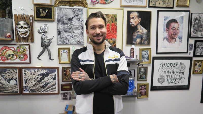 En leende man med mörkt kort hår, bruna ögon och kort helskägg står med armarna i kors framför en vägg fullhängd med foton på tatueringar.