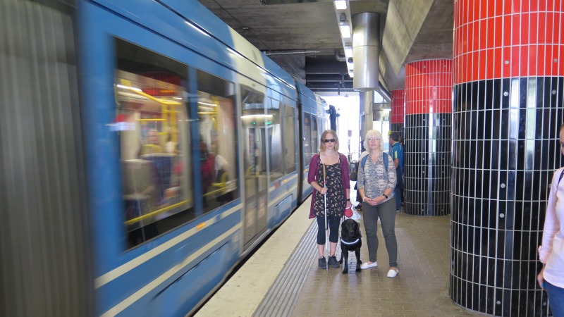 En kvinna med mörka glasögon, vit käpp och ledarhund vid sin sida står intill en blond, korthårig kvinna på en perrong medan ett tåg kommer körande till vänster i bild.