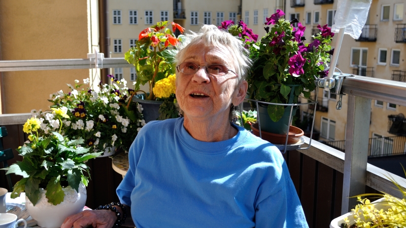 Barbro Lindell har kort grått hår, glasögon och en blå tröja. Hon är omgiven av blommarna på sin balkong.