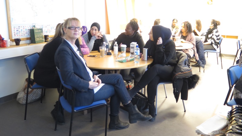 Läraren Cecilia Lindeberg vid ett runt bord i ett klassrum. I bakgrunden studenter inbegripna i livligt samtal.
