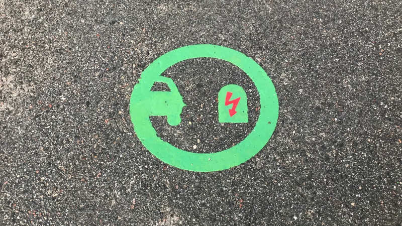 Symbol på asfalt i en parkeringsficka för laddning av elbilar. I en neongrön cirkel syns en halv bilkaross till vänster och en laddningstation med röd elblixt till höger.