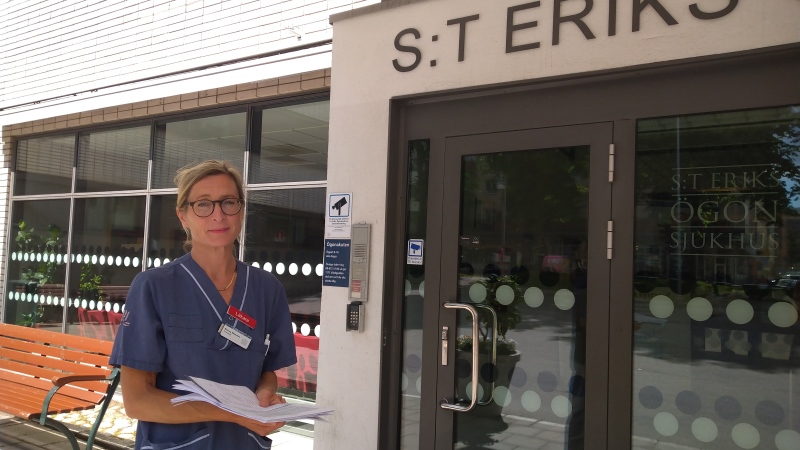 Blond kvinna med håret samlat i tofs, bruna glasögon och klädd i blå sjukhuskläder framför sjukhusets huvudingång.