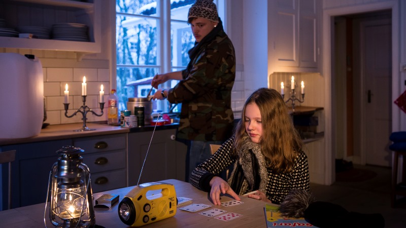 Interiör från ett kök utan el. En man i vinterkläder lagar mat på spritkök, en flicka spelar kort vid bordet. Flera ljusstakar brinner och på bordet står en fotogenlampa och en radio.