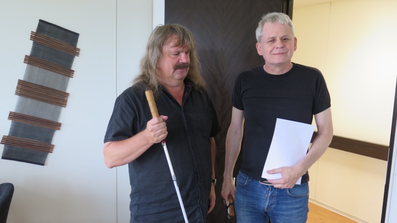 Håkan Thomson har axellångt hår och mustasch och håller i en vit käpp, Lennart Karlsson har kort, grått hår och håller i ett papper på punkt i ena handen och en ihopvikt käpp i den andra. De står i dörröppningen till en korridor.