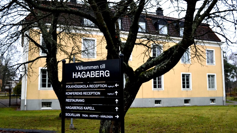 Stort gult hus i puts, en skola, ligger i en park. I förgrunden ett avlövat träd och en stor skylt för Hagabergs folkhögskola.