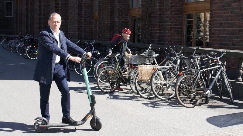 Daniel Helldén, en vithårig man i blå kostym utan slips står med ena foten på en elsparkcykel som han är på väg att parkera.