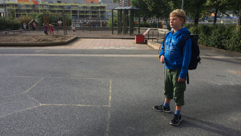 En pojke står med vit käpp och ryggsäck på en skolgård.