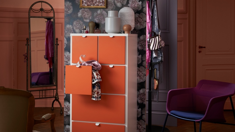 Foto ur Ikeas katalog. Del av färgglatt rum med fokus på en hög, vit byrå med stora orange lådor framför en storblommig tapet. Mönstrade kläder på en klädhängare och en lila fotölj intill.