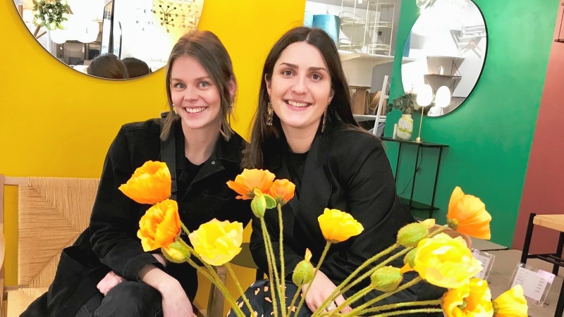 Två unga kvinnor sitter bredvid varandra, knallgul vägg bakom, stor bukett gula blommor i vas framför.