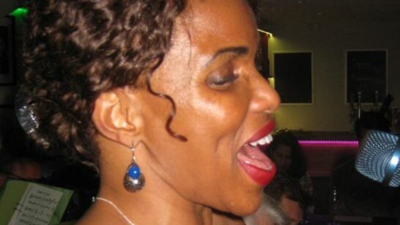 Kvinna med mörk hy och uppsatt lockigt hår sedd i profil framför mikrofon.