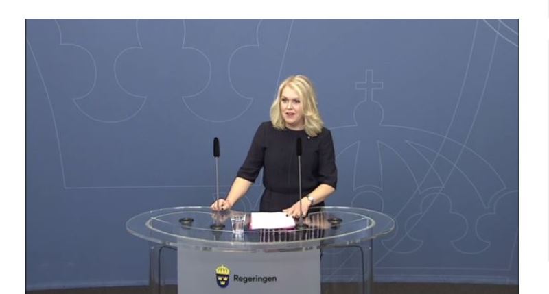 Lena Hallengren har halvlångt blond hår och står vid ett talarbord med mikrofoner.