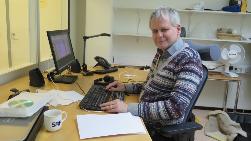 Lennart Karlsson, ombudsman för SRF. Han har kort, ljusgrått hår och randig tröja och sitter vid sitt skrivbord framför datorn. På bordet finns också en hög med punktskrift och en kaffekopp.