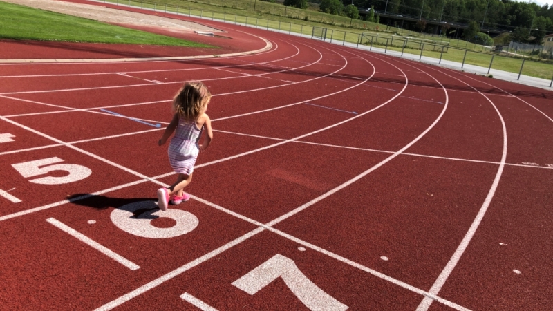 En liten blond flicka i ljus sommarklänning springer på en roströd löparbana.