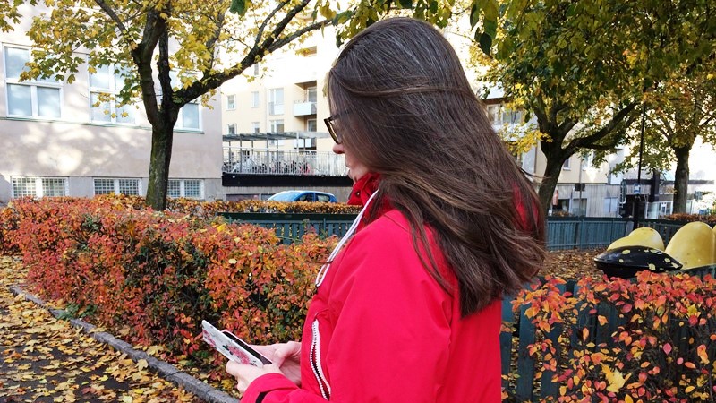 Bild bakifrån: Kvinna med mörkt hår och röd jacka, går och tittar ner på mobilen. Träd i bakgrunden.