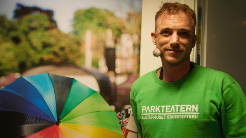 Till höger i bild står Albin Flinkas i en ljusgrön tröja med texten Parkteatern. Han är kortklippt med rufsigt hår och skäggstubb. Till vänster i bild finns en plansch med ett regnbågsfärgat paraply.
