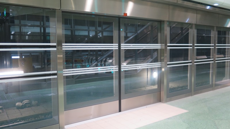 Bild på plattformsbarriärer vid perrongkanten, en vägg av glas med vita horistontala streckmarkeringar.