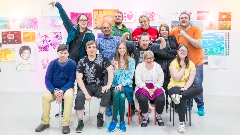 Lars Lerin och Junior med tio av deltagarna som medverkar i Lerins lärlingar. Alla skrattar och är samlade framför en vägg med färgglada målningar. Foto Ulrika Malm SVT legal