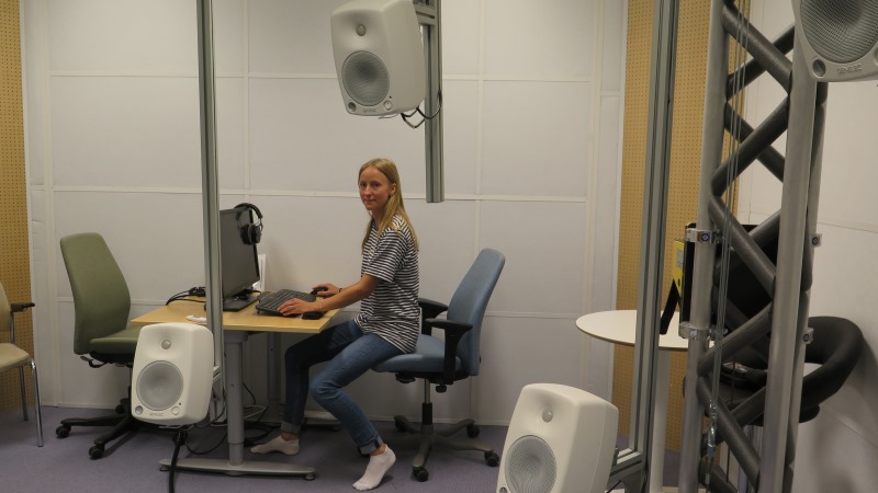 Kvinna med långt blont hår och randig tröja sitter vid en dator i en ljudstudio med flera små högtalare som hänger på ställningar.
